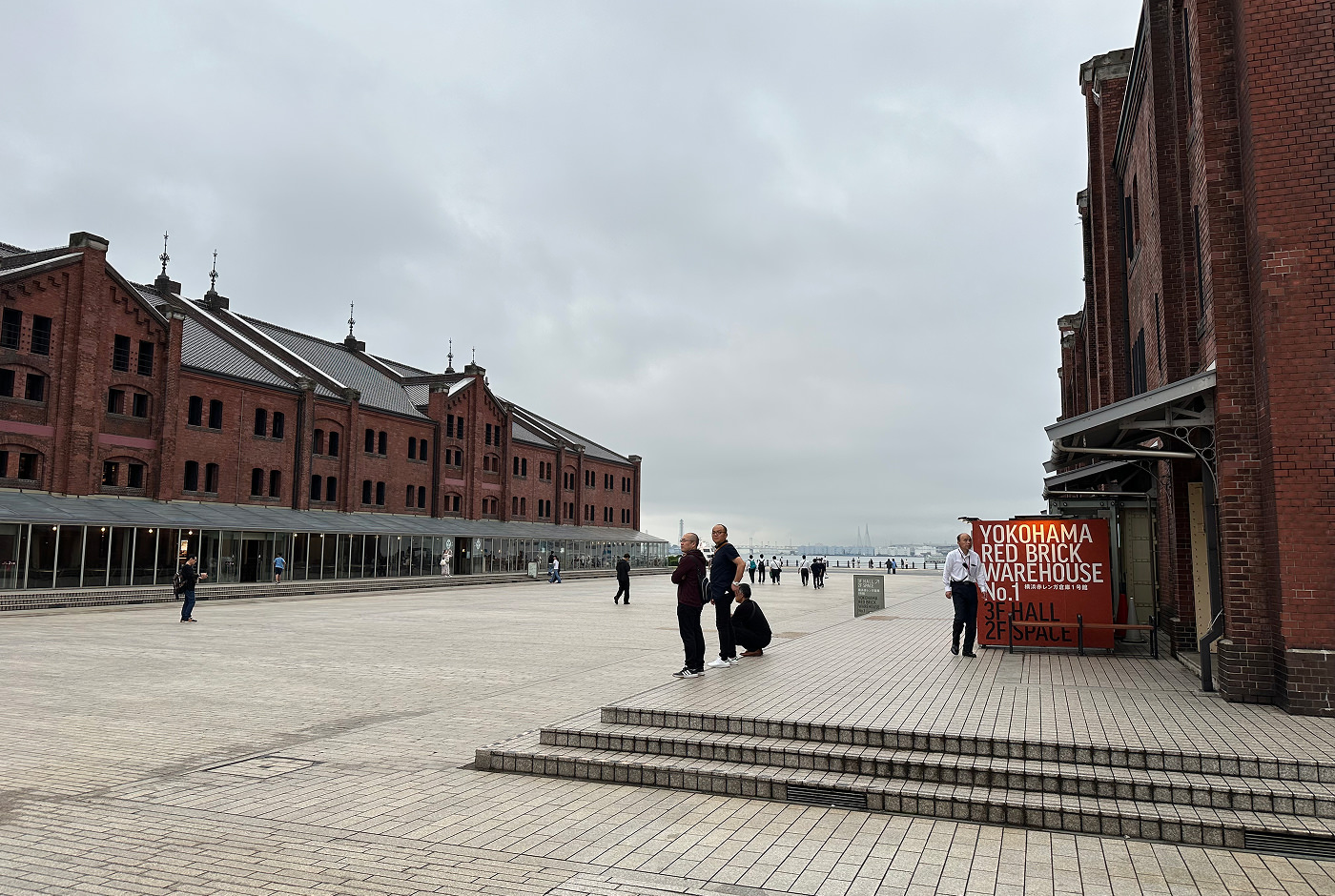 Två enorma hus i rött tegel står på varsin sida om ett stort öppet ljust torg. Ett tiotal personer går på torget.