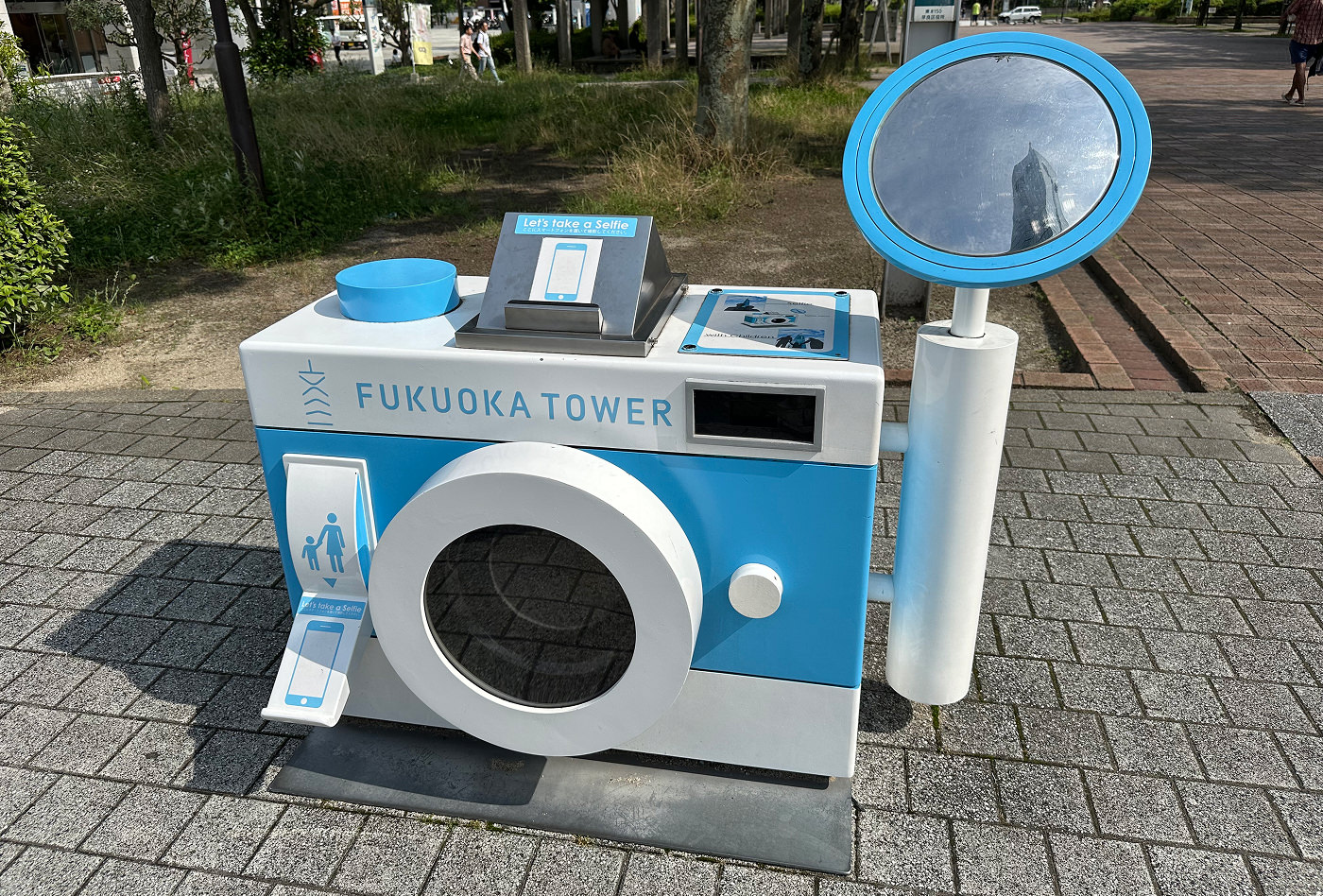 En blå och vit skulptur av en klassisk kamera i en park med text som lyder "Fukuoka Tower". En spegle är monterad på sidan.