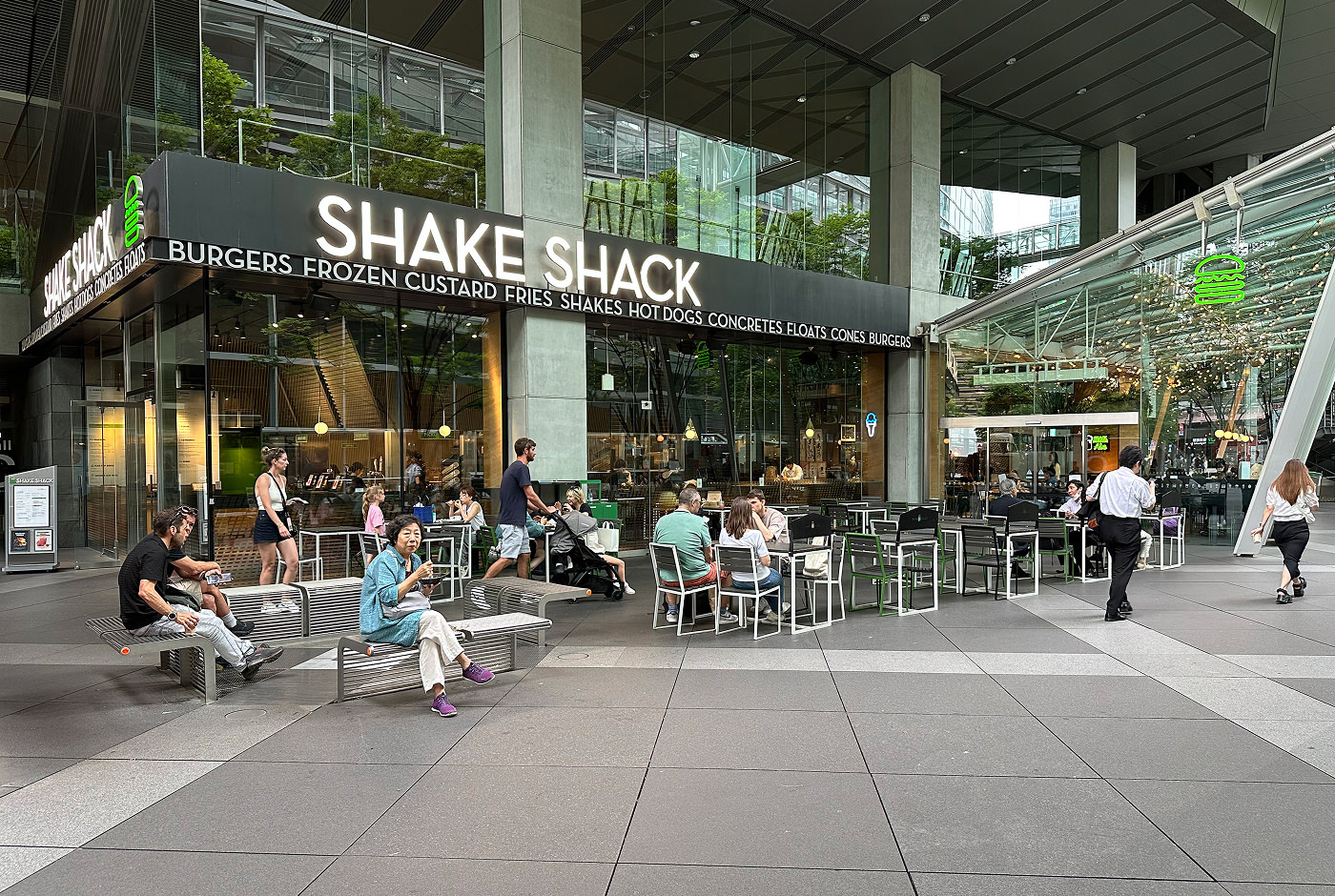 Glasfasaden för en Shack Shack-restaurang. Folk sitter både inne och ute och äter. I bakgrundens syns en stor byggnad som också har glasfasad.
