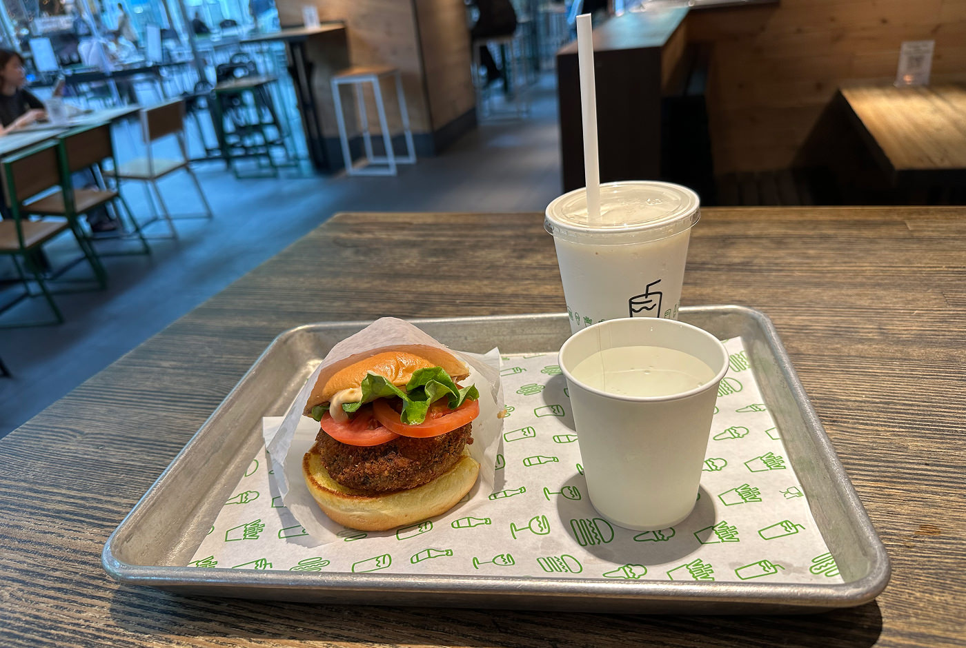 Ljus plåtbricka med en hamburgare och två vita papperskoppar, en med vatten och en med milkshake.