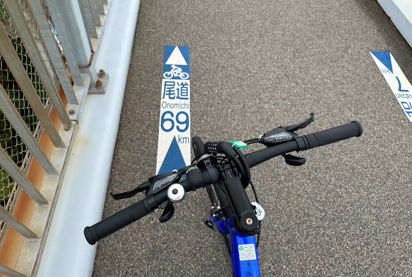 Styre på en cykel. Nedanför är ett märke målat på vägen med texten "Onomichi 69 km".