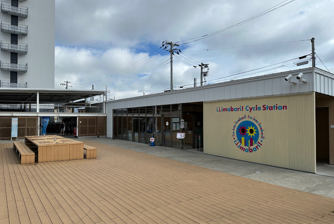 Stor yta med ljus trägolv mellan flera byggnader. En skylt lyder "Imabari Cycle Station".