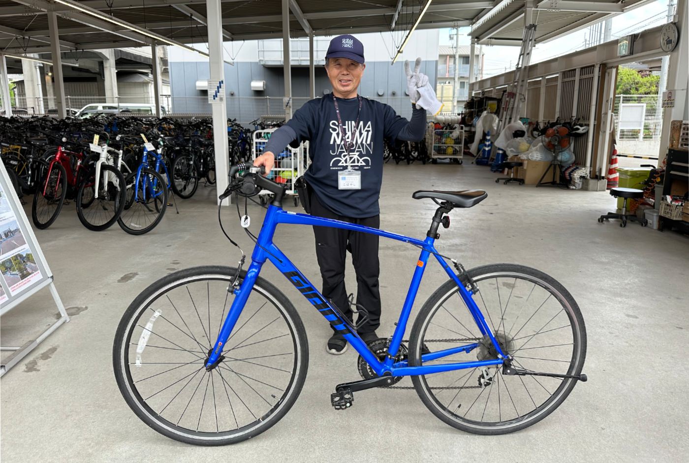 Äldre japansk man i mröka kläder håller i en blå cykel, ler och visar fredstecknet.