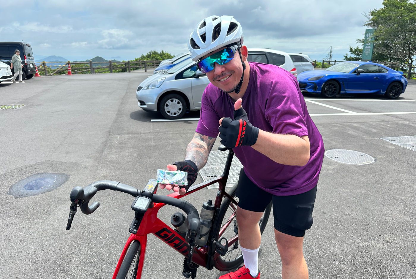 Japansk cyklist med lila tröja och vit hjälm ler och håller i en kaka och ger tummen upp.