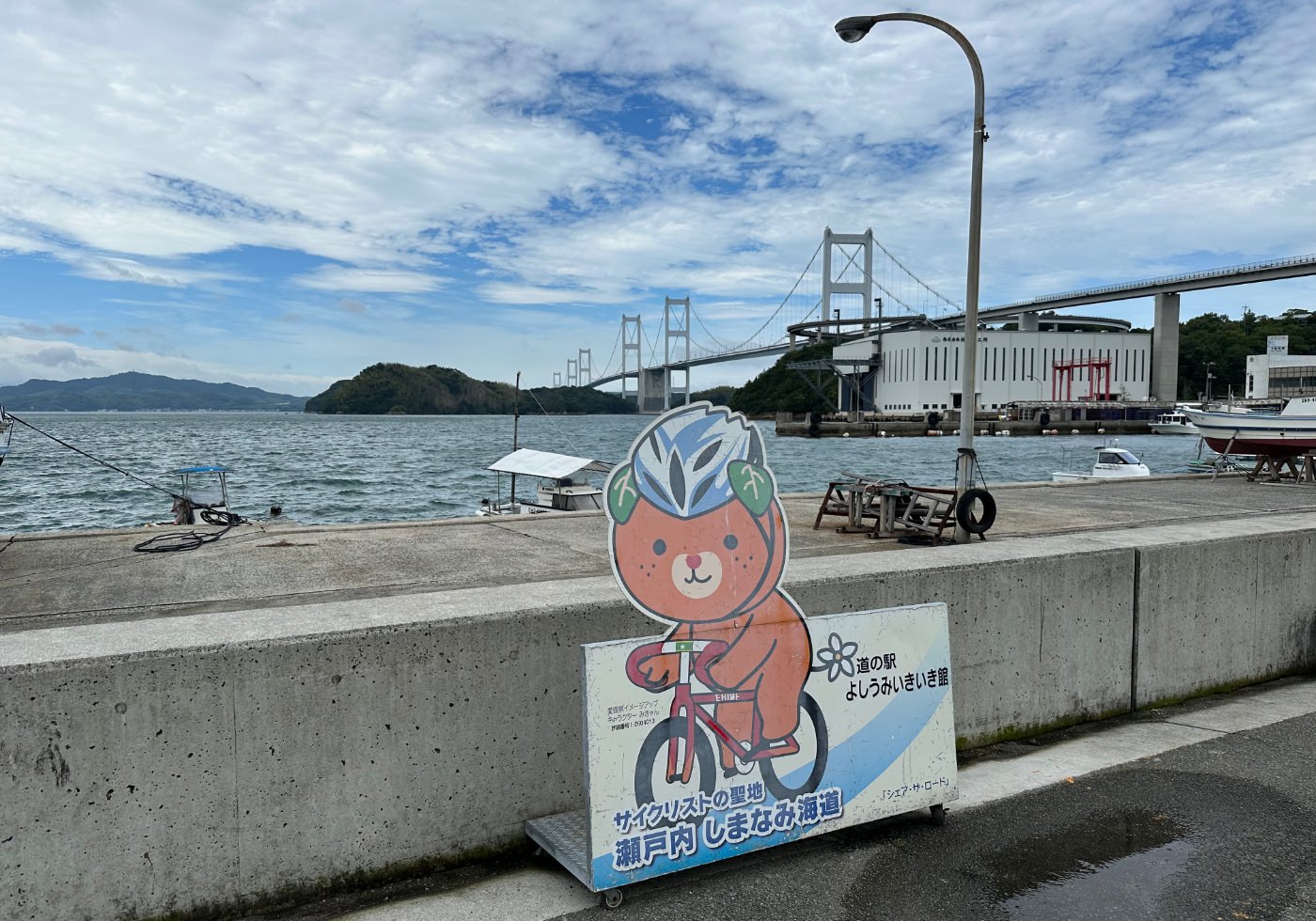 Reklamskylt med en orange maskot som cyklar. I fjärran en kilometerlång hängbro över ett inlandshav.