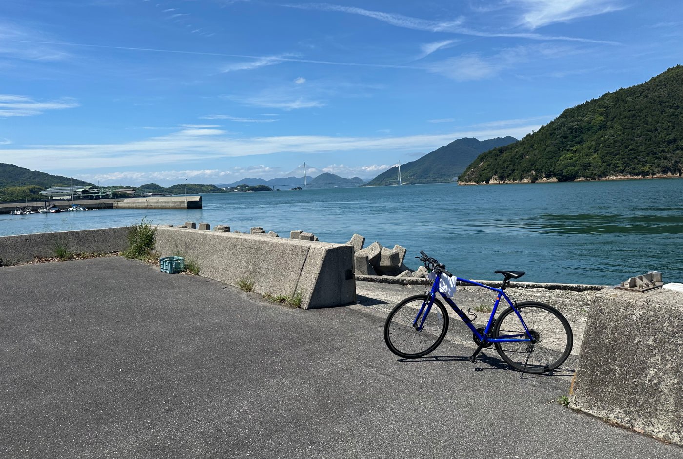 En blå cykel står parkerad vid en betongpir. I fjärran lummiga berg och en lång vit bro mellan två öar.