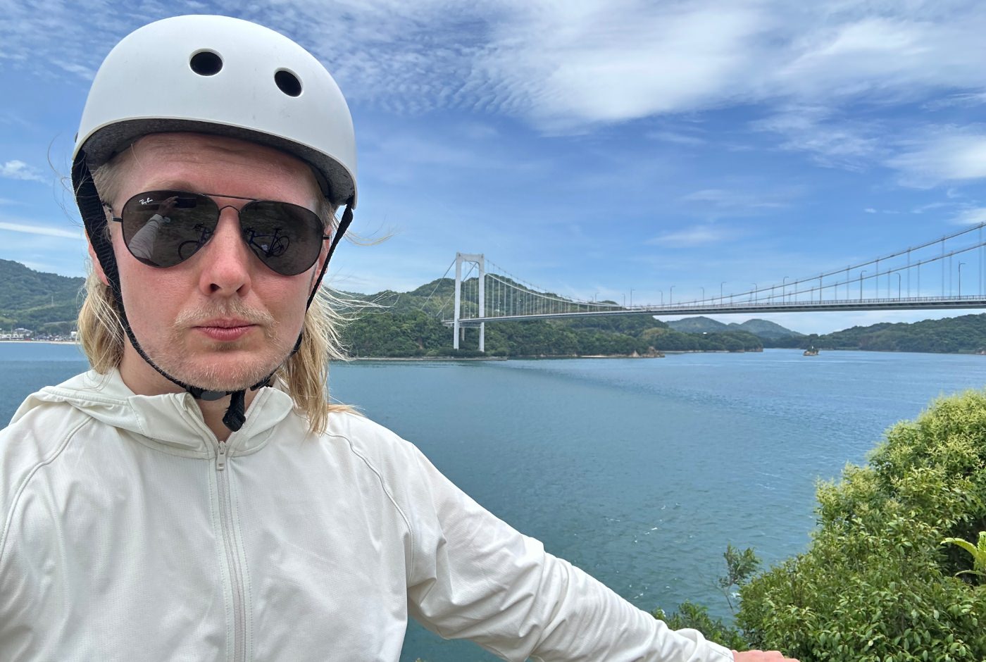 Jag har vit jacka, vit cykelhjälm, solglasögon och tar en selfie. Bakom mig är vit bro mellan två öar i ett hav.
