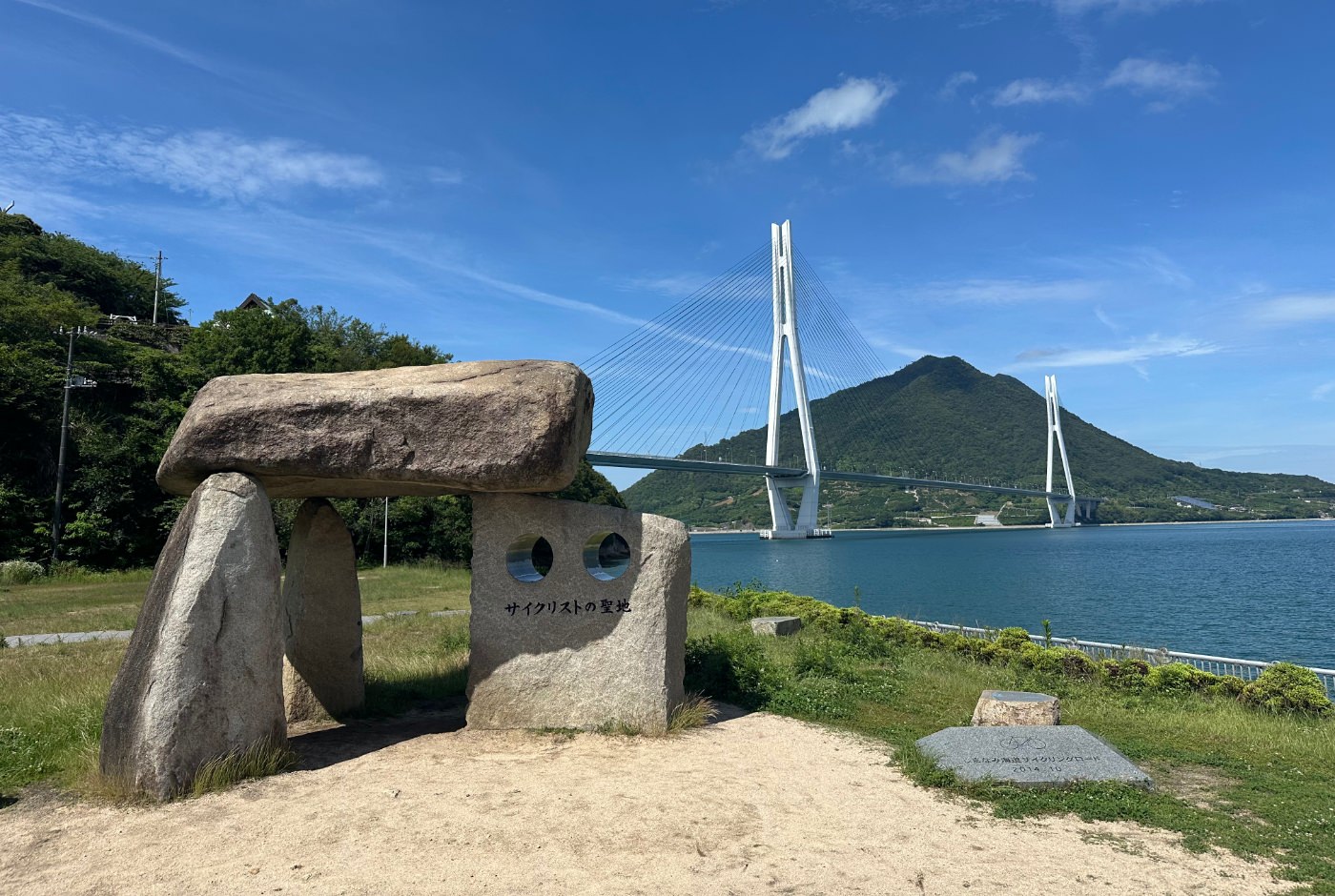 En skulptur med fyra stenar framför en stor vit bro över havet mellan två lummiga berg.