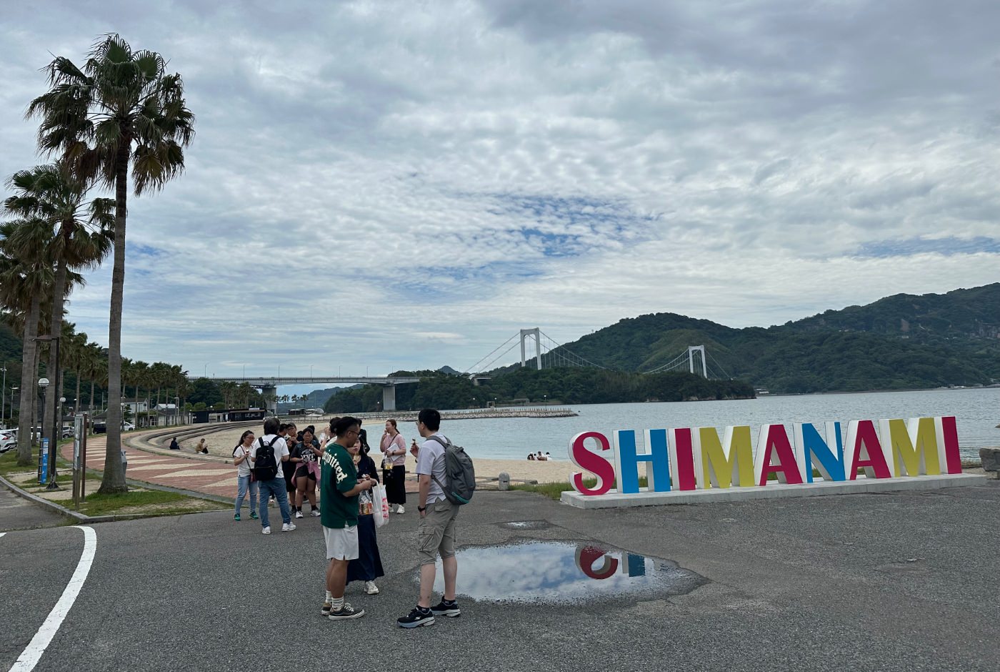 En grupp människor står framför en böjd sandstrand med palmer. En stor skylt lyder "Shimanami". I fjärran lummiga berg och en bro.
