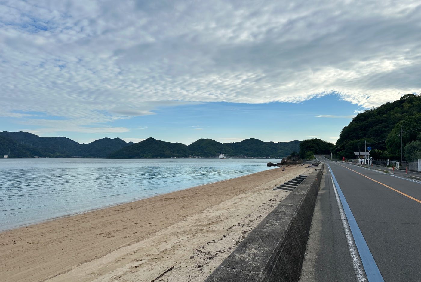 Bil- och cykelväg längs med en sandstrand. På andra sidan finns en lummig bergskedja.
