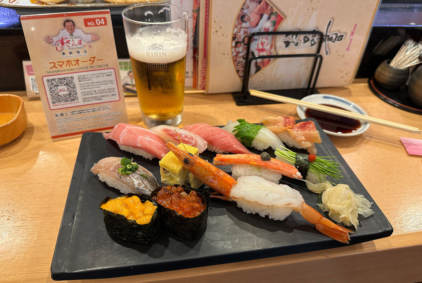 Svart bricka med tolv bitar sushi. Bakom den på trädisken står ett glas öl.