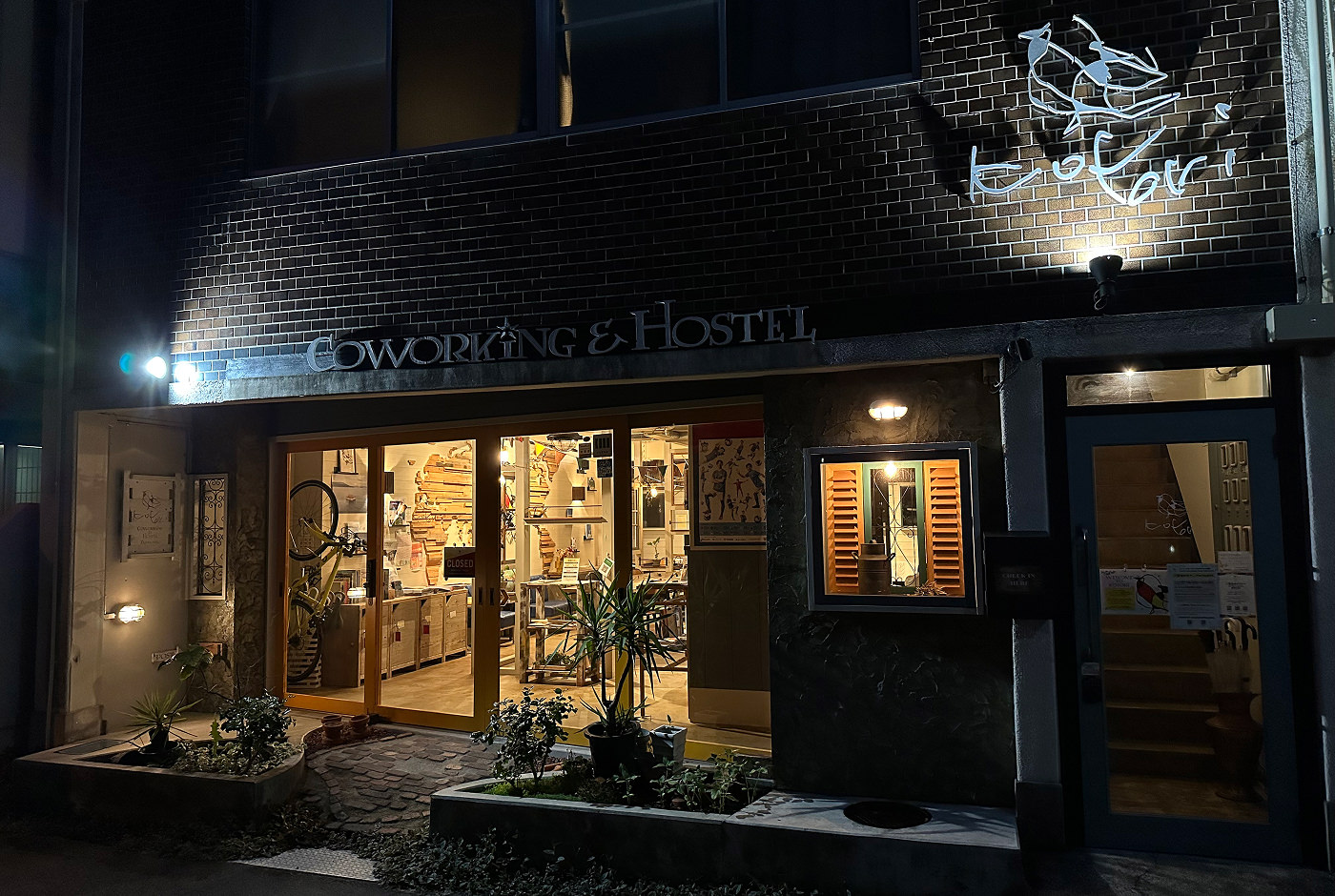 Upplyst fasad för ett vandrarhem sent på kvällen. Man ser in i lobbyn genom flera glasdörrar. Skylten lyder "Kotori Coworking & Hostel".