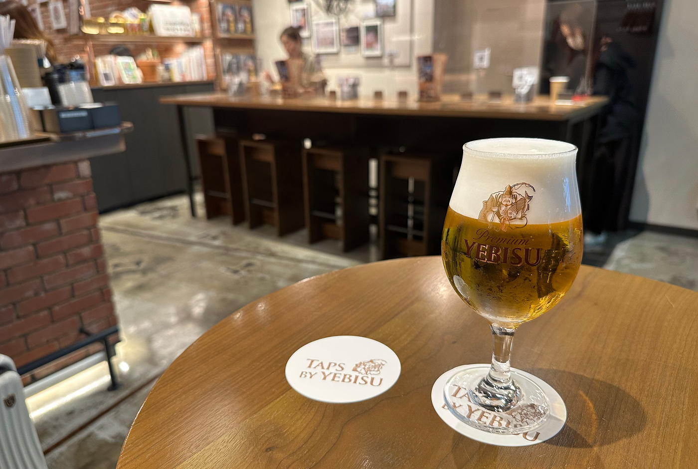 Ett runt glas öl står på ett runt träbord. Ett vitt drinkunderlägg har texten "Taps by Yebisu".