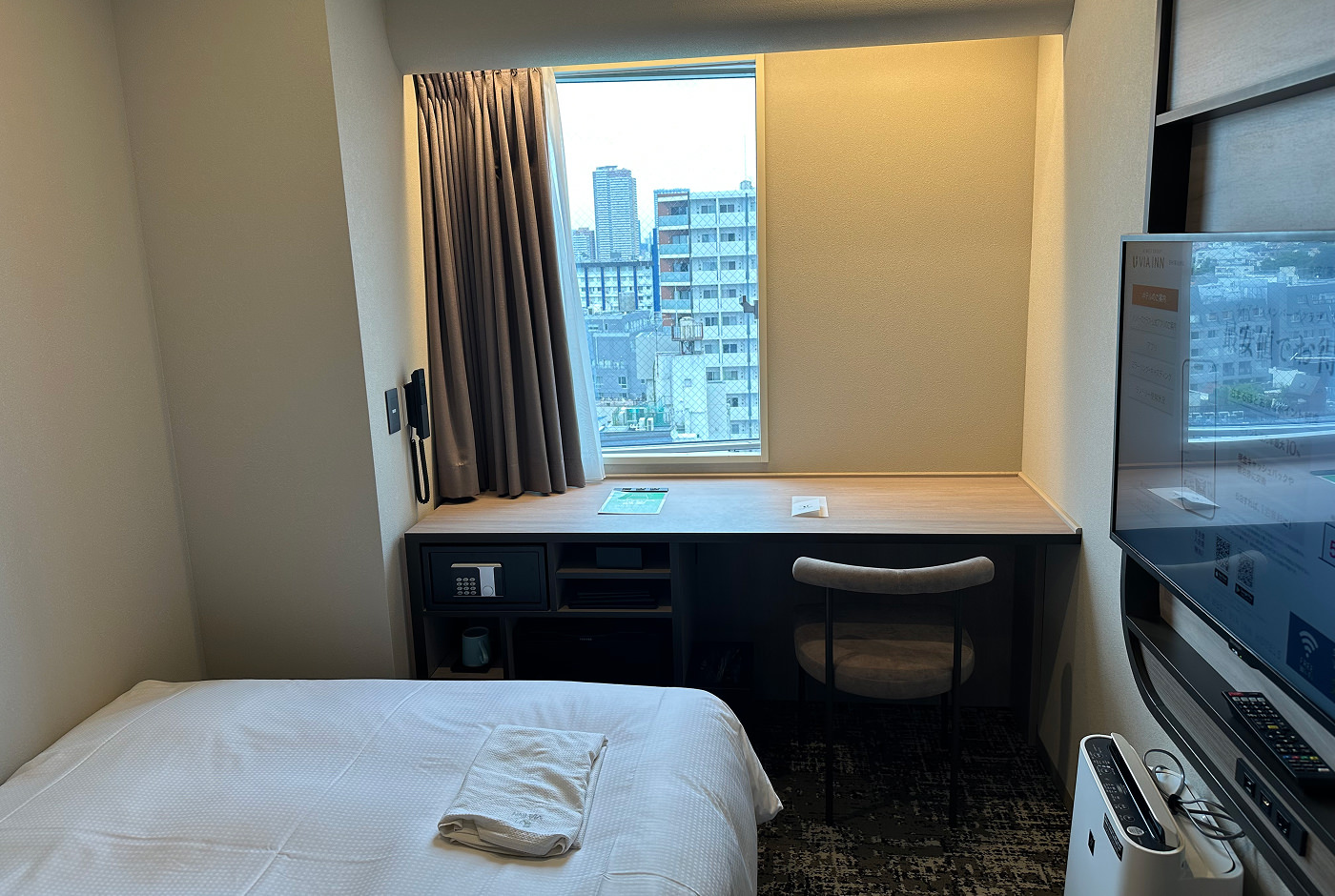 Ljust hotellrum med litet fönster ovanför ett skrivbord.