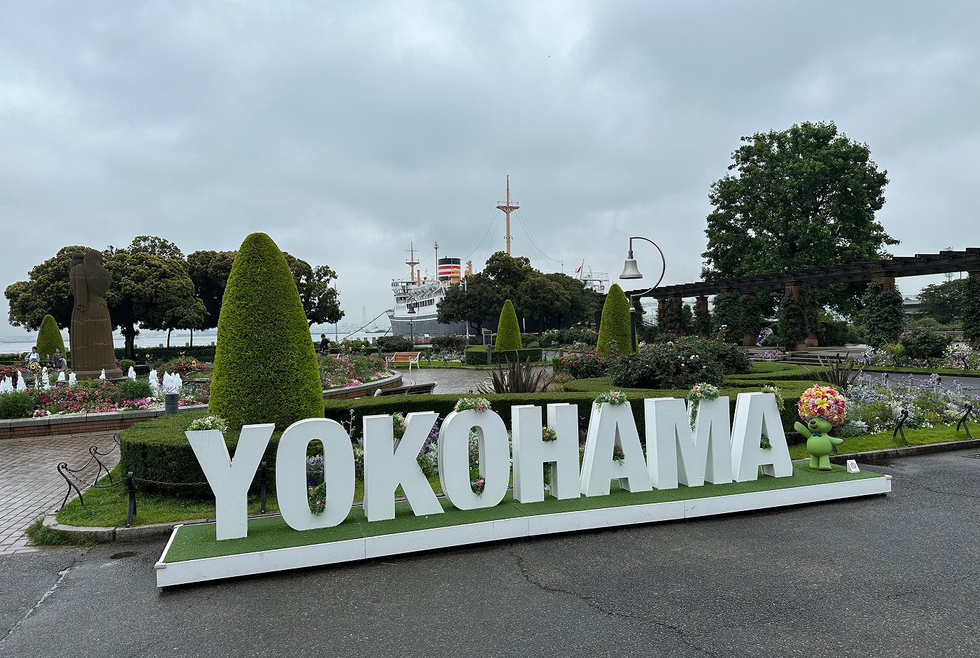 En vit skylt som lyder "Yokohama" står framför en park och ett ankrat fartyg.