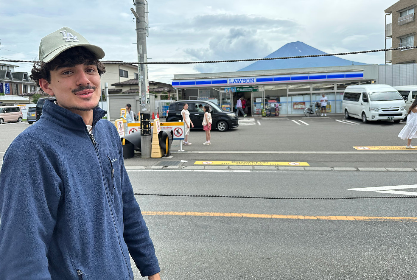 Ung kille med blå tröja och ljus keps ler in i kameran framför en liten matbutik framför Mount Fuji.