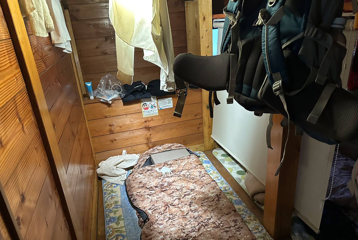 Liten sovplats i ett vandrarhem. På golvet ligger en sovsäck och på en hylla ovanför den ligger lite kläder. I förgrunden hänger en ryggsäck från taket.