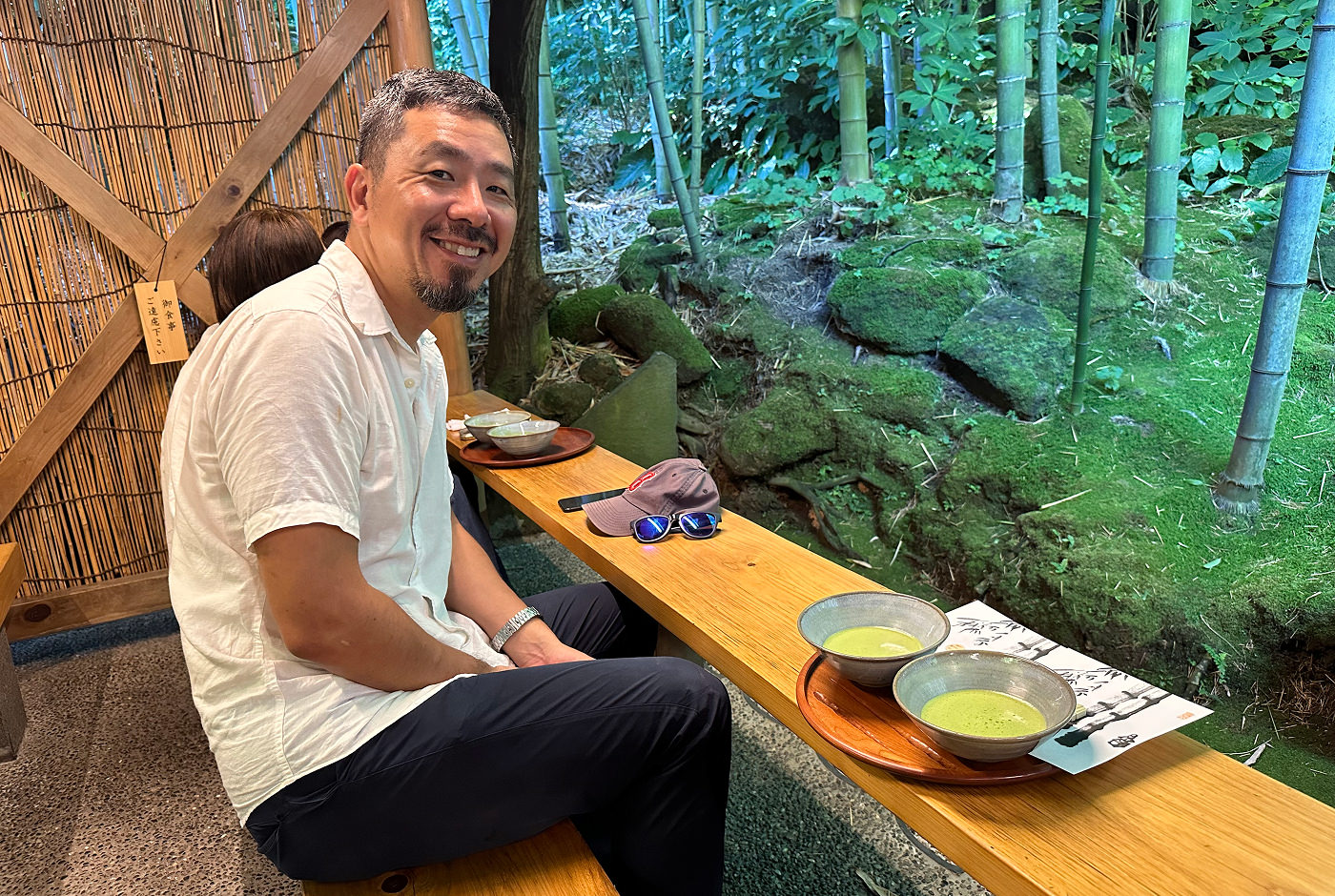 Japansk man med vit skjorta och svarta byxor sitter på en träbänk vid en skog och ler i i kameran. På bänken står två skålar med matchate.