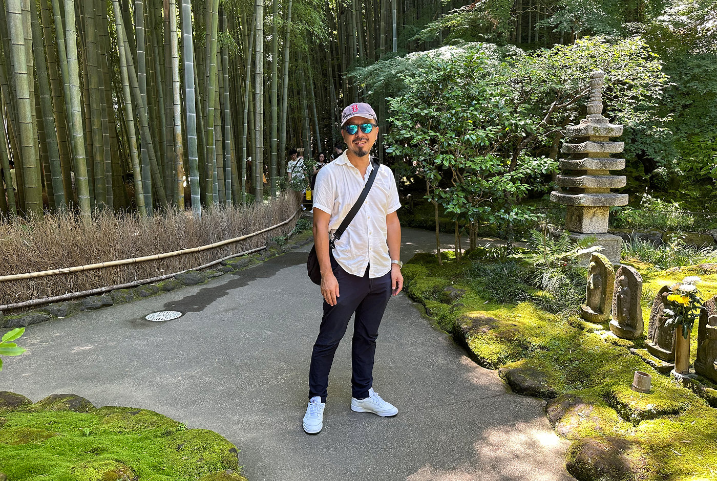 Japansk man med svarta byxor, vit skjorta, blå keps och solglasögon står vid en stengång vid en bambuskog.