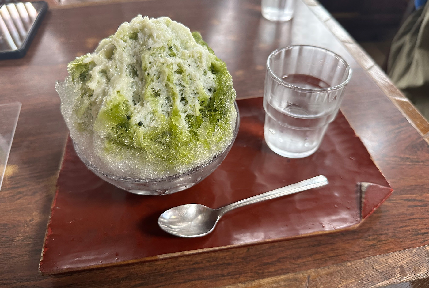 Mörkröd träbricka på ett bord. På brickan står ett glas vatten, en sked och en skål med skivad is toppad med grön matcha.