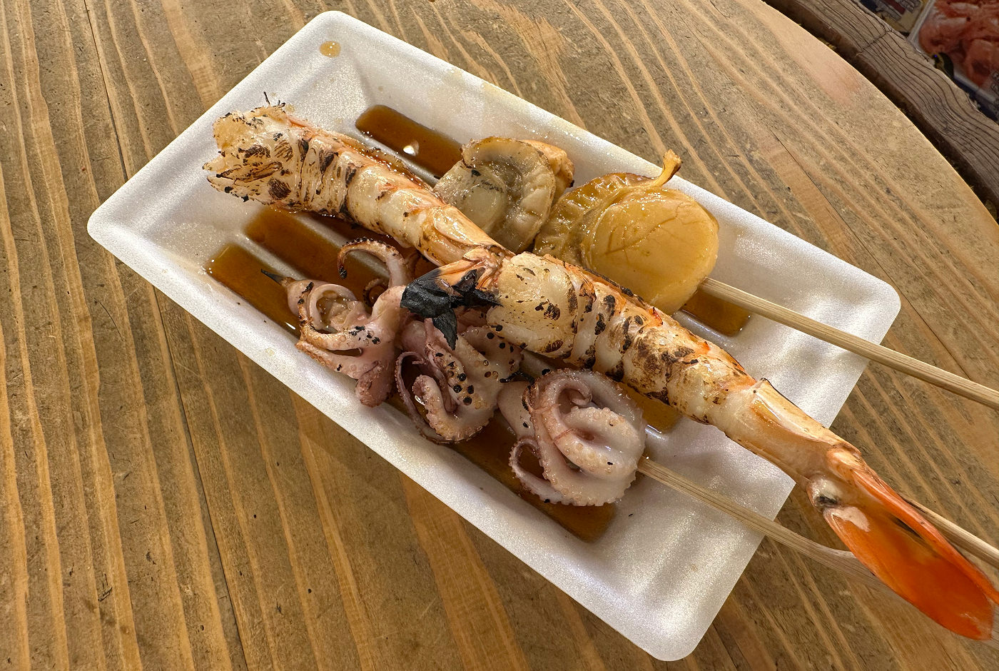 Vit papperstallrik med flera grillspett med bläckfisk, jätteräkor och kammusslor.