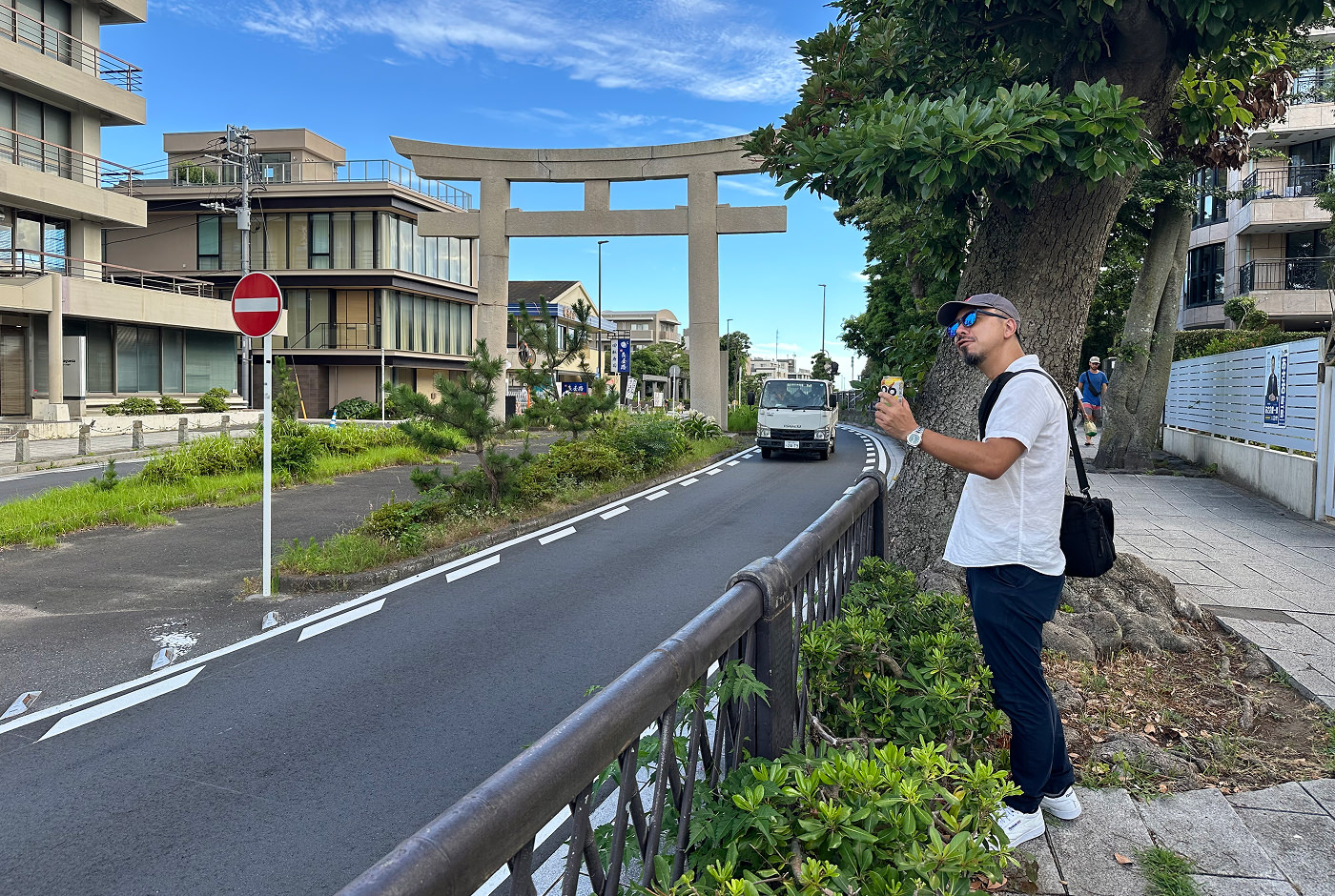 Japansk med med svarta byxor, vit skjorta, keps och solglasögon håller i en ciderburk vid sidan av en väg som klyvs av en torii-port.