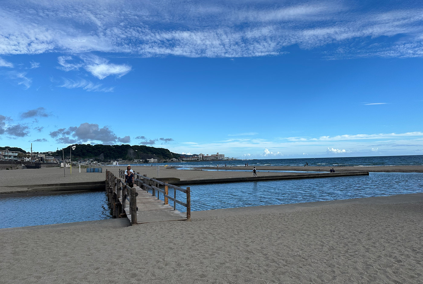Stor sandstrand med en träbrygga över en liten kanal som delar stranden. I fjärran vid kusten ser man lummiga berg med bebyggelse vid kusten.