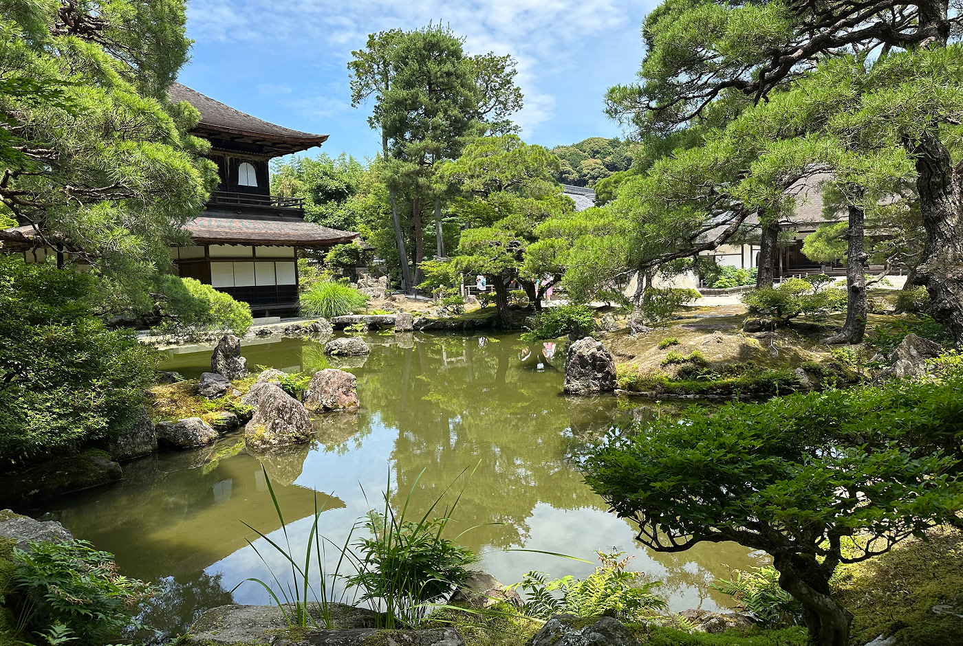 Liten damm i en traditionell japansk landskapsträdgård. Till vänster syns en träpaviljong med två våningar. Till höger kan man se flera byggnader genom träden.