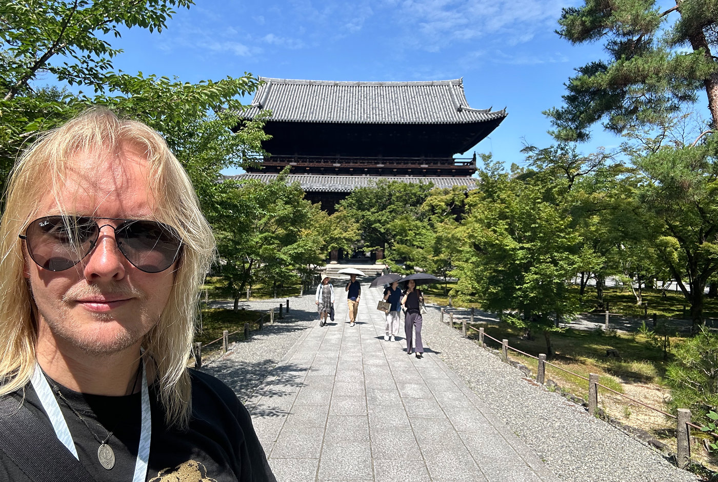Jag har solglasögon på mig och tittar leende in i kameran. Bakom mig står ett stort gammalt japanskt tempel med böjt tak och två våningar. En ljus stengång till templet är omgiven av buskiga träd.