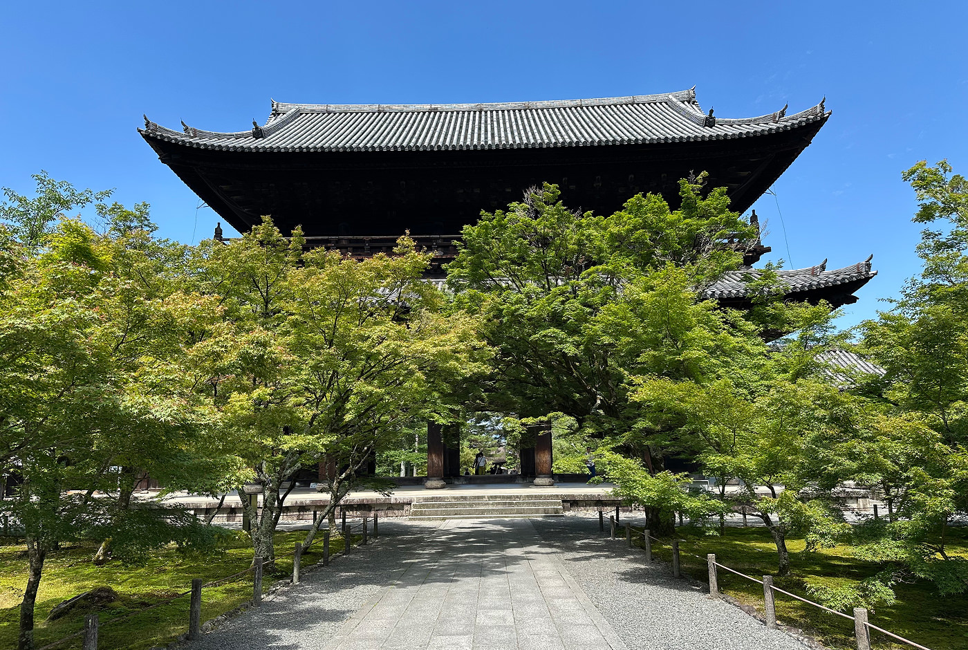 Stort gammalt japanskt tempel med böjt tak och två våningar. En ljus stengång till templet är omgiven av buskiga träd.