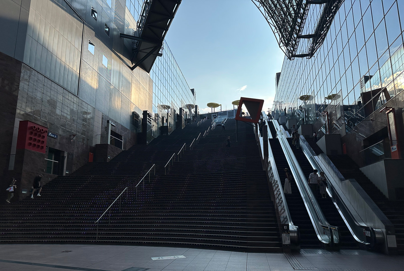 En mycket bred och lång trappa i flera sektioner som leder upp till ett område under bar himmel på en tågstation. Till höger finns även två rulltrappor.