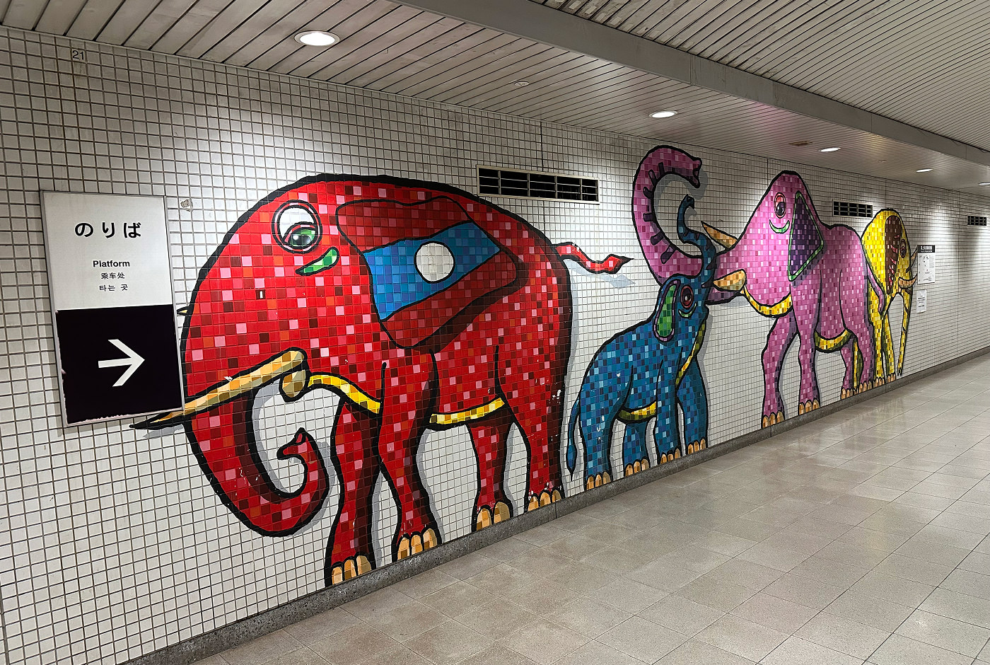 Vit kakelvägg i en tunnelbanestation. Fyra karikatyrer på på elefanter är målade på kaklet. En röd, blå, rosa och gul.