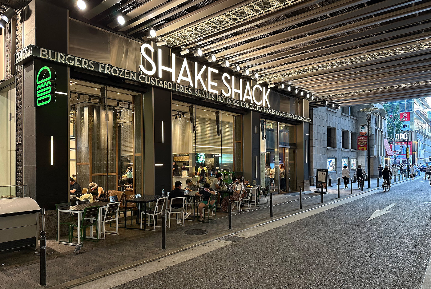 Upplyst glasfasad till hamburgarkedjan Shake Shack. Fasaden ligger under en bro vid en enkelriktad väg.