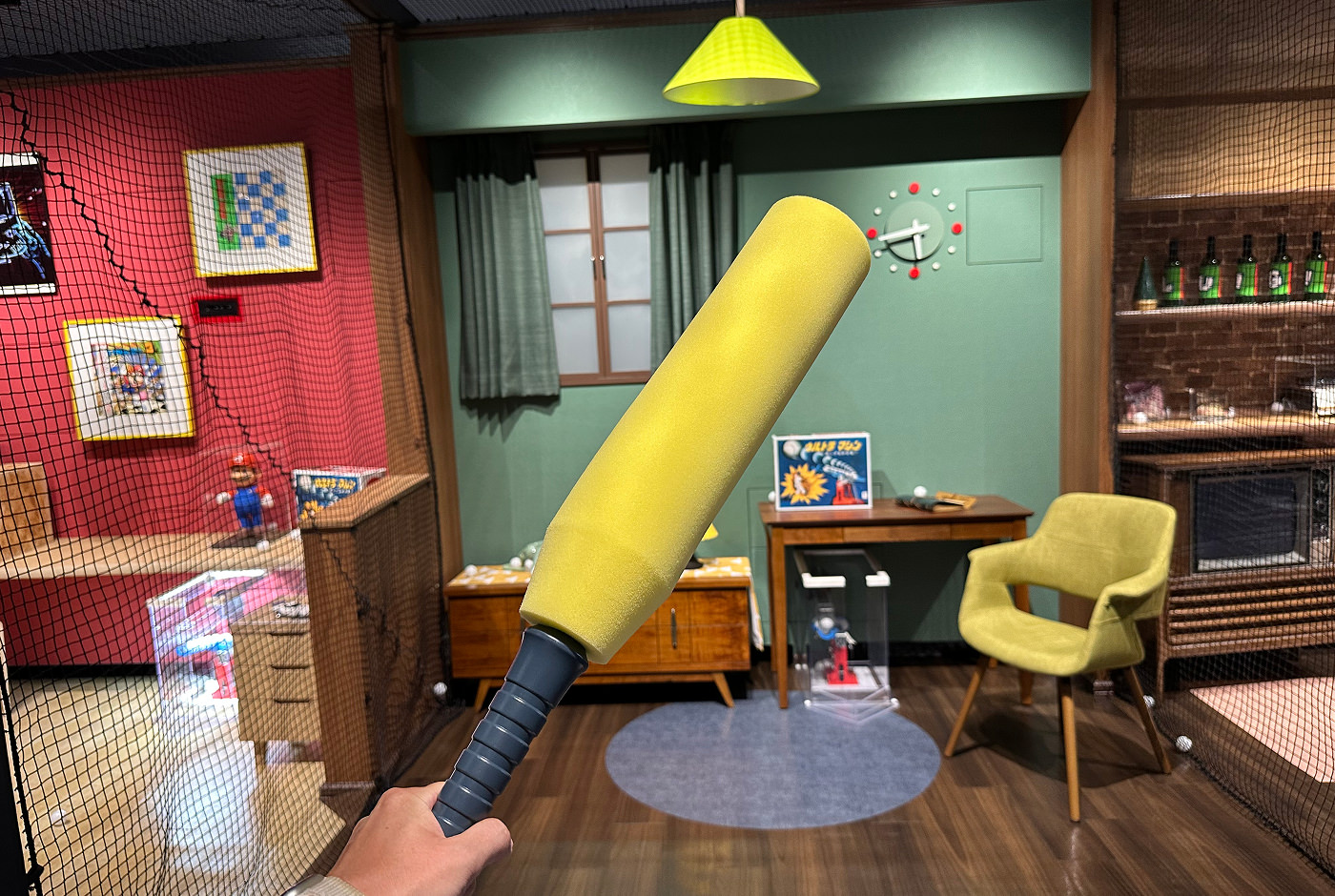 Hand håller i ett basebollträ i skumgummi. I bakgrunden ser man en lekfull tolkning av ett rum i ett egnahemshus.