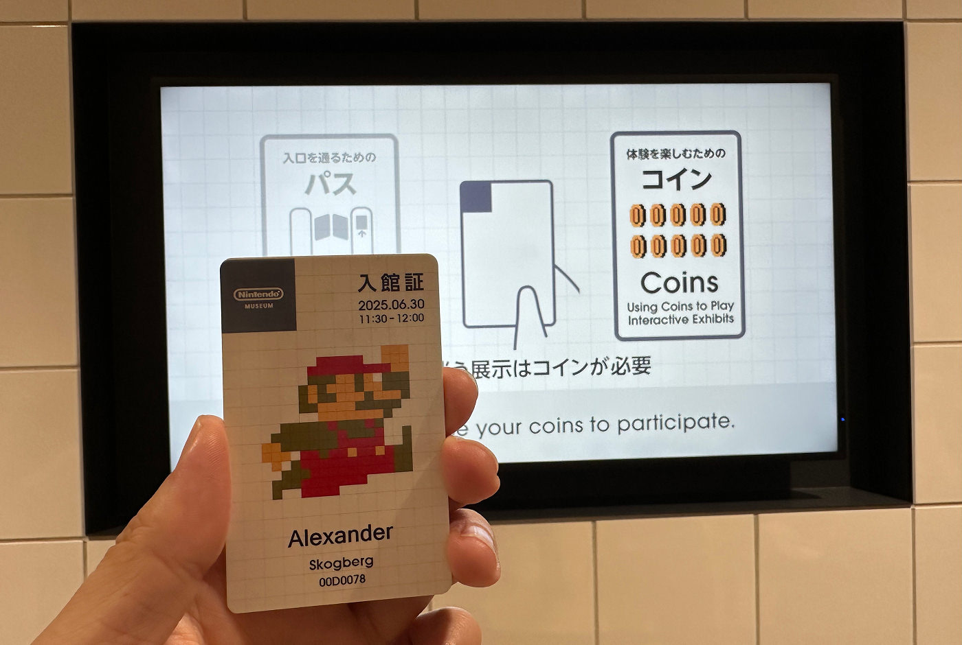 Hand gåller i en vit plastbiljett framför en skärm med instruktioner hur den fungerar. Biljetten har en bild på Super Mario och texten "Alexander Skogberg".