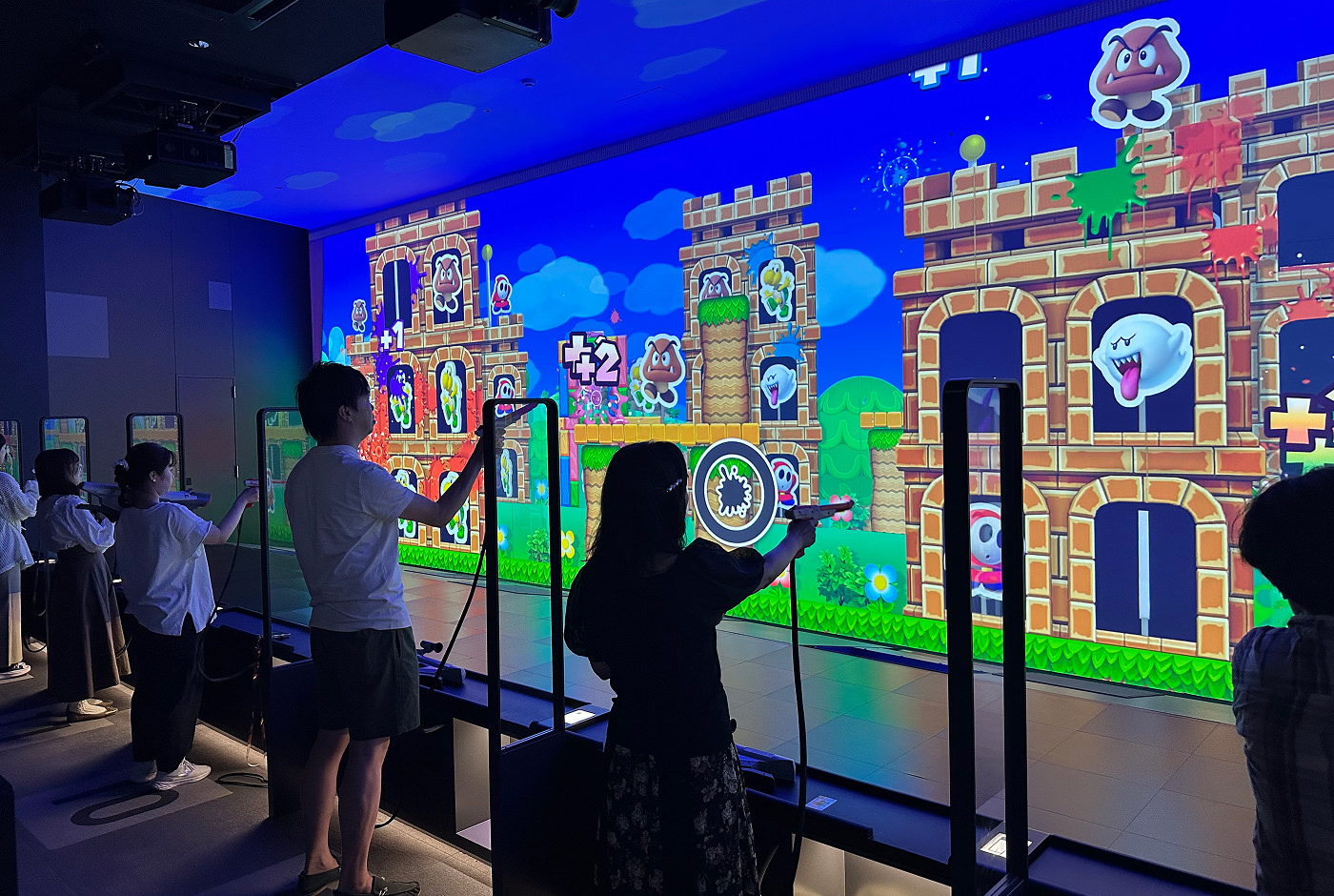Enormt bred skärm fylld med fiender från Super Mario-spelen. Framför står flera personer med plastpistoler och skjuter digitala färgkulor mot dessa fiender.
