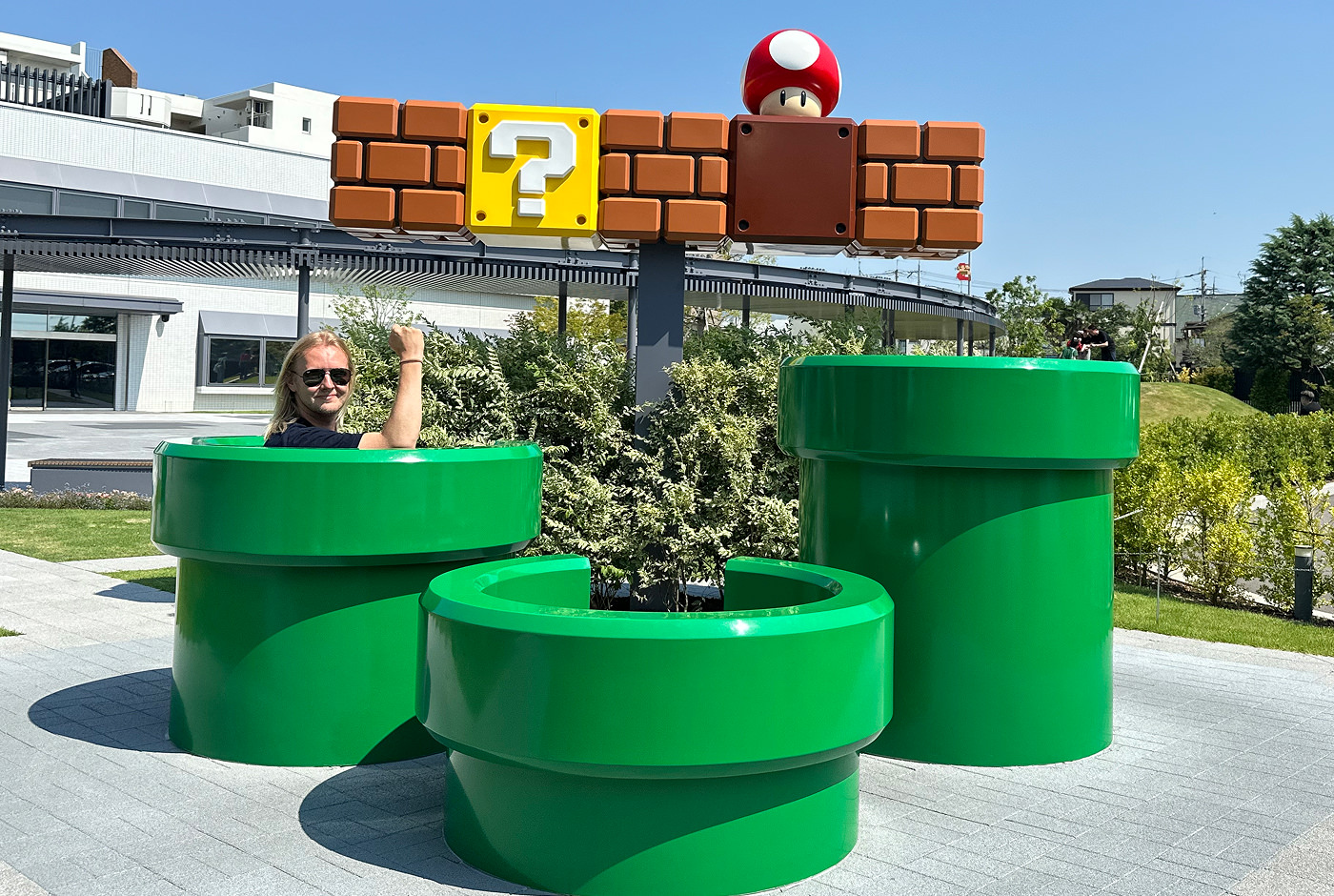 Installation med tre gröna rör, tre tegelblock och två Power Up-block från Super Mario spelen. Jag sitter i ena röret och lyfter högernäven upp mot skyn.