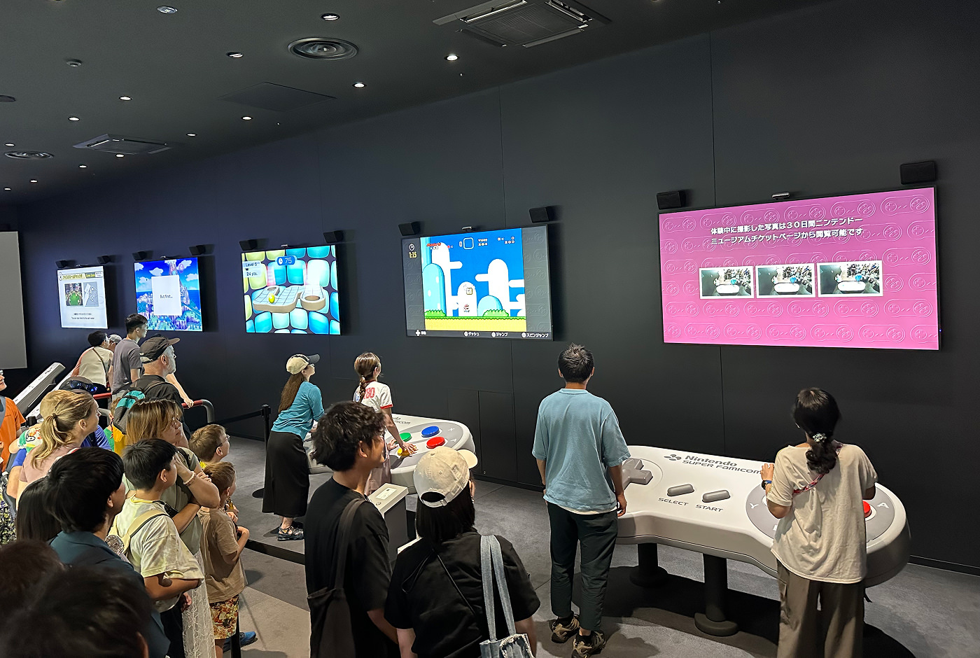 Stort rum där folk spelar Nintendos klassiska spel på breda skärmar. Två personer står framför enorma kontrollers och samarbetar för att spela.