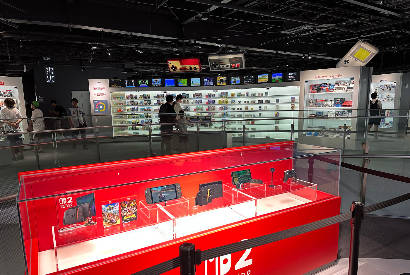 Röd monter som visar Nintendos nya bärbara spelkonsol Switch 2. I bakgrunden sons flera ljusa och upplysta glasmontrar med spel från förr i tiden. Ett tiotal personer står och tittar på montrarna