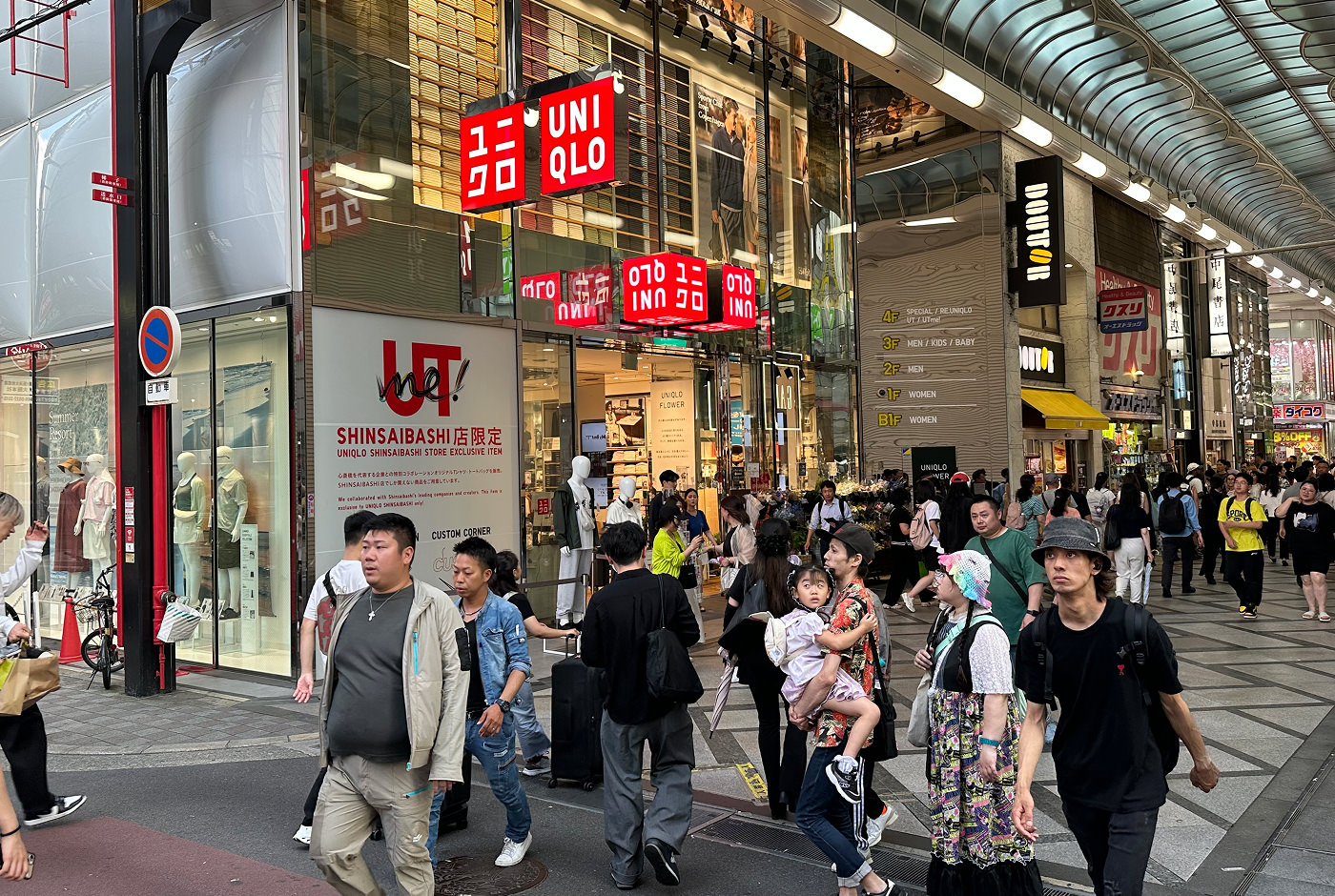 Bred shoppingarkad fylld med folk och butiker med färggranna skyltar. Dne närmaste butiken är Uniqlo med sina rödvita skyltar. En har texten "Shinsaibashi".