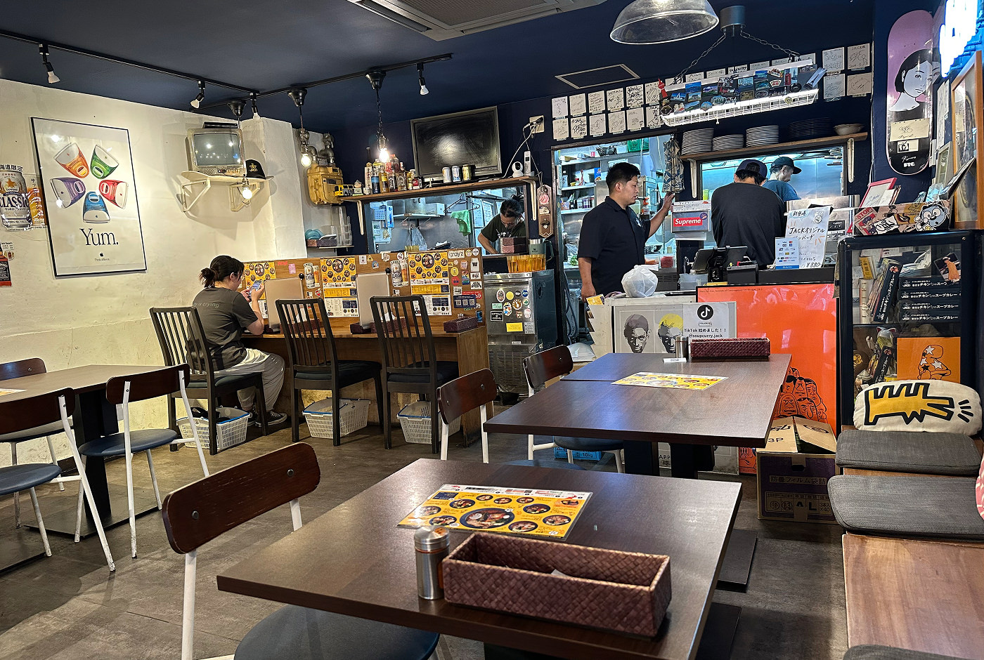 Restaurang med halvöppet kök och flera bruna bord. Fyra personer står i köket och en person sitter och äter. På väggarna finns flera färggranna planscher.