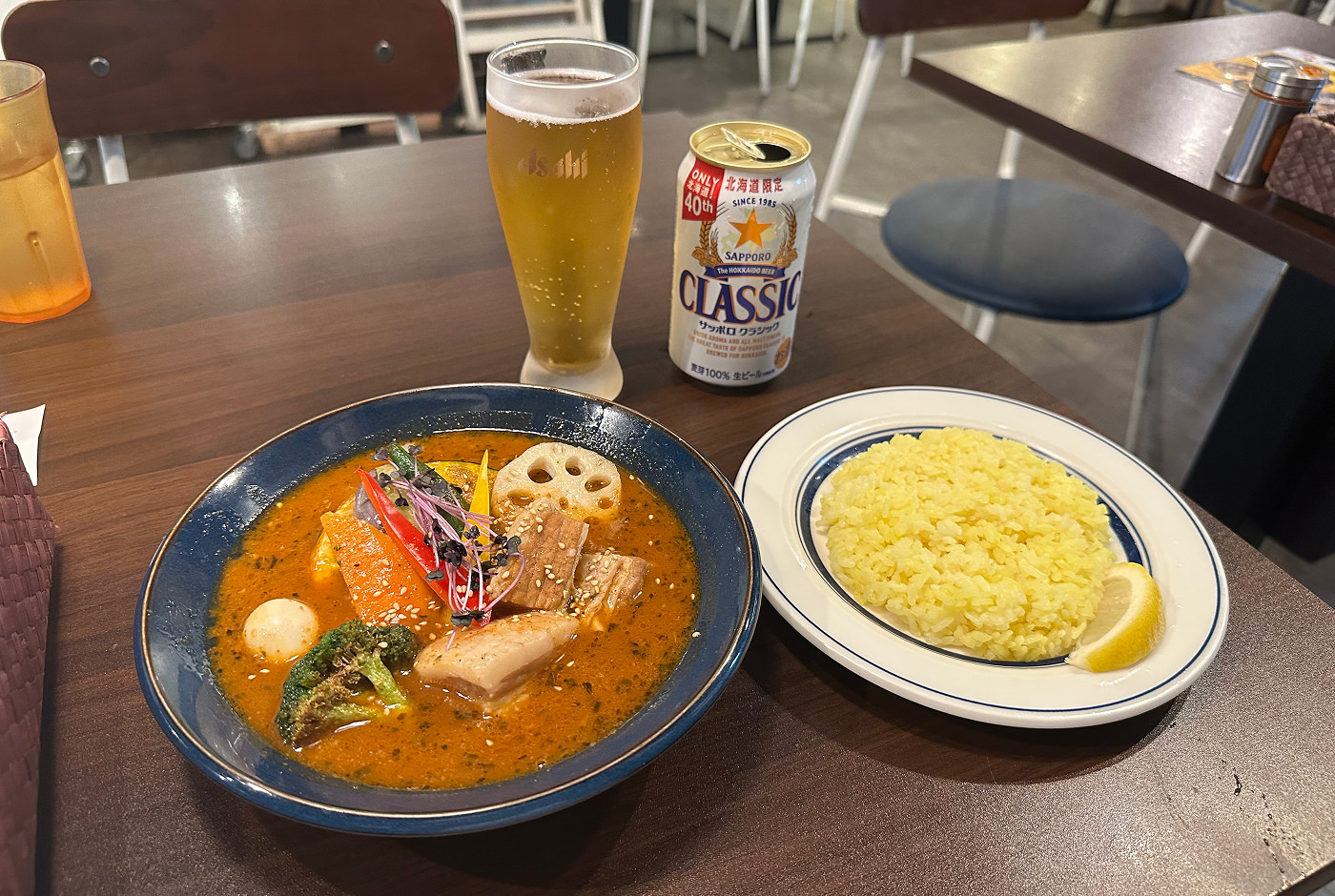 En mörk skål med orange currysoppa med grönsaker står på ett bord bredvid en vit tallrik med ris och ett upphällt ölglas.