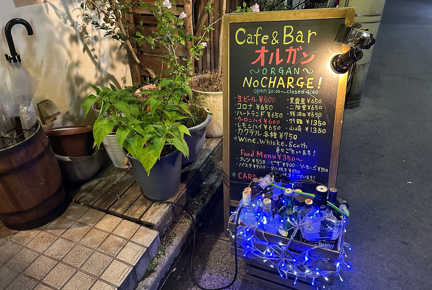 Svart griffeltavla står på marken jämte flera blommor i vaser. Text på tavlan lyder "Café & Bar Organ".
