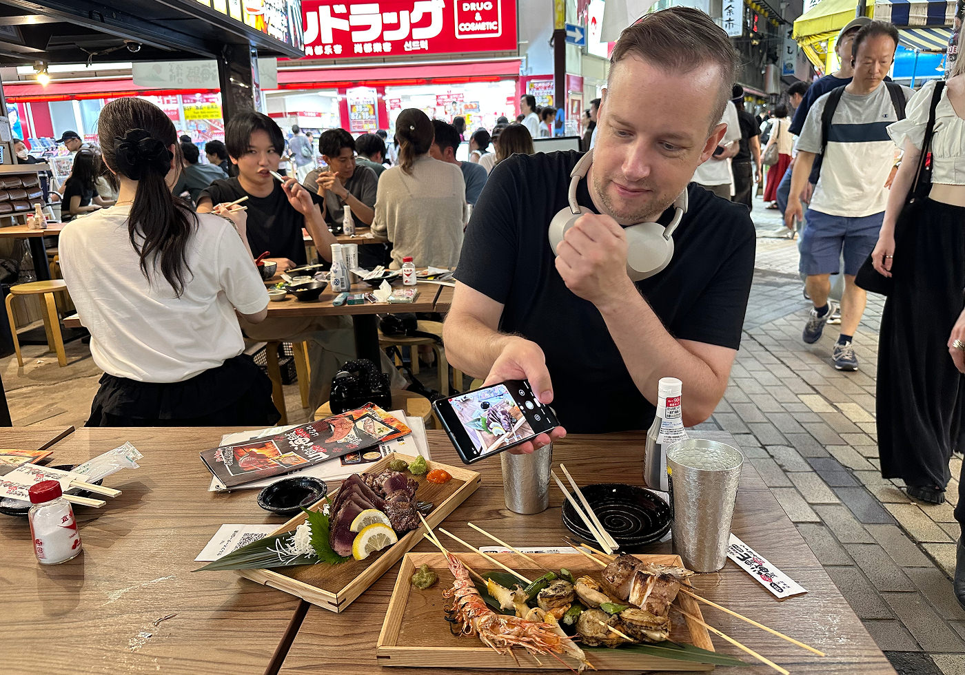 Dan sitter vid ett träbord på en uteservering och fotograferar två träbrickor med grillspett och fisk.