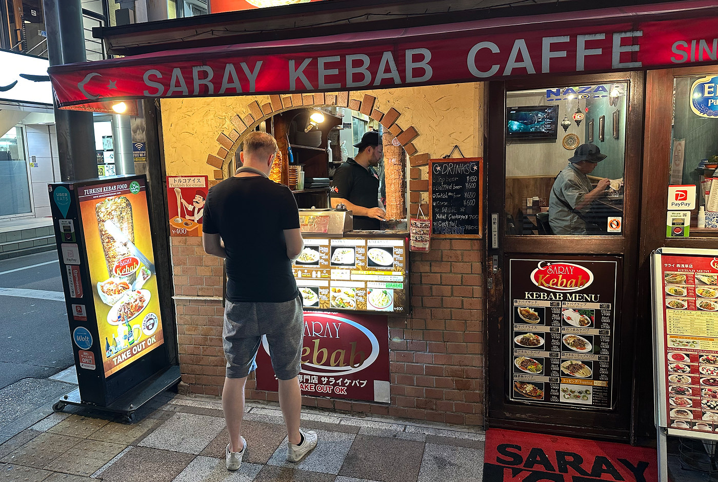 Man med gråa shorts och mörk t-shirt står vid ett litet upplyst kebabhak och beställer mat. Hakets skylt lyder "Saray Kebab Caffe".