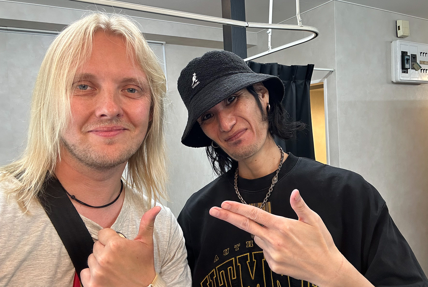 Jag står bredvid en japansk man med svart hatt, svart tröja och långt svart hår. Vi ler båda in i kameran och pekar på varandra.