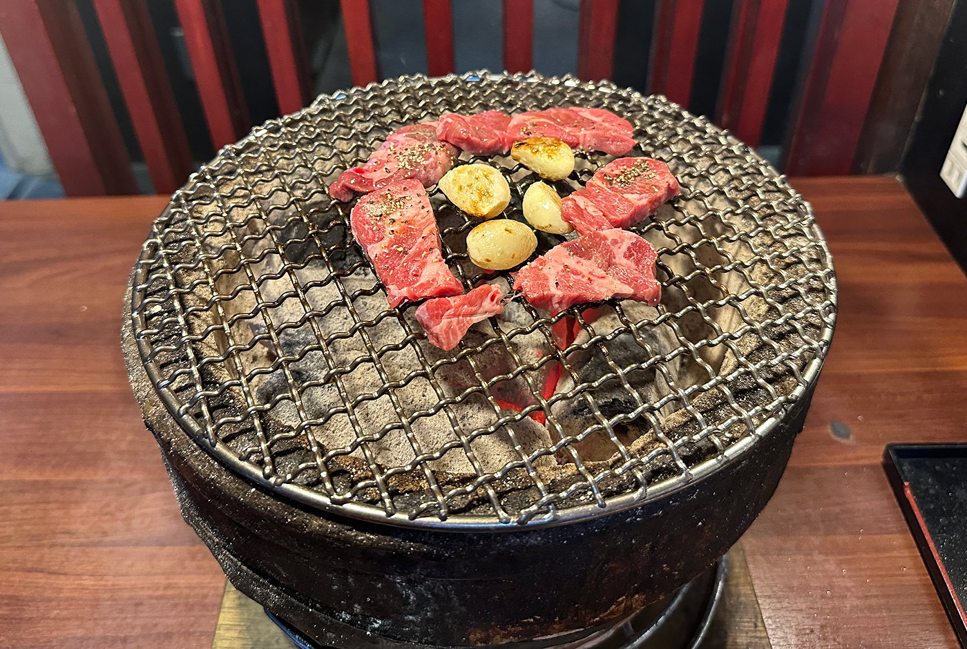 En liten kolgrill av sten på ett bord. Den har ett stålgaller ovanpå med fem små köttstycken och fyra vitlöksklyftor