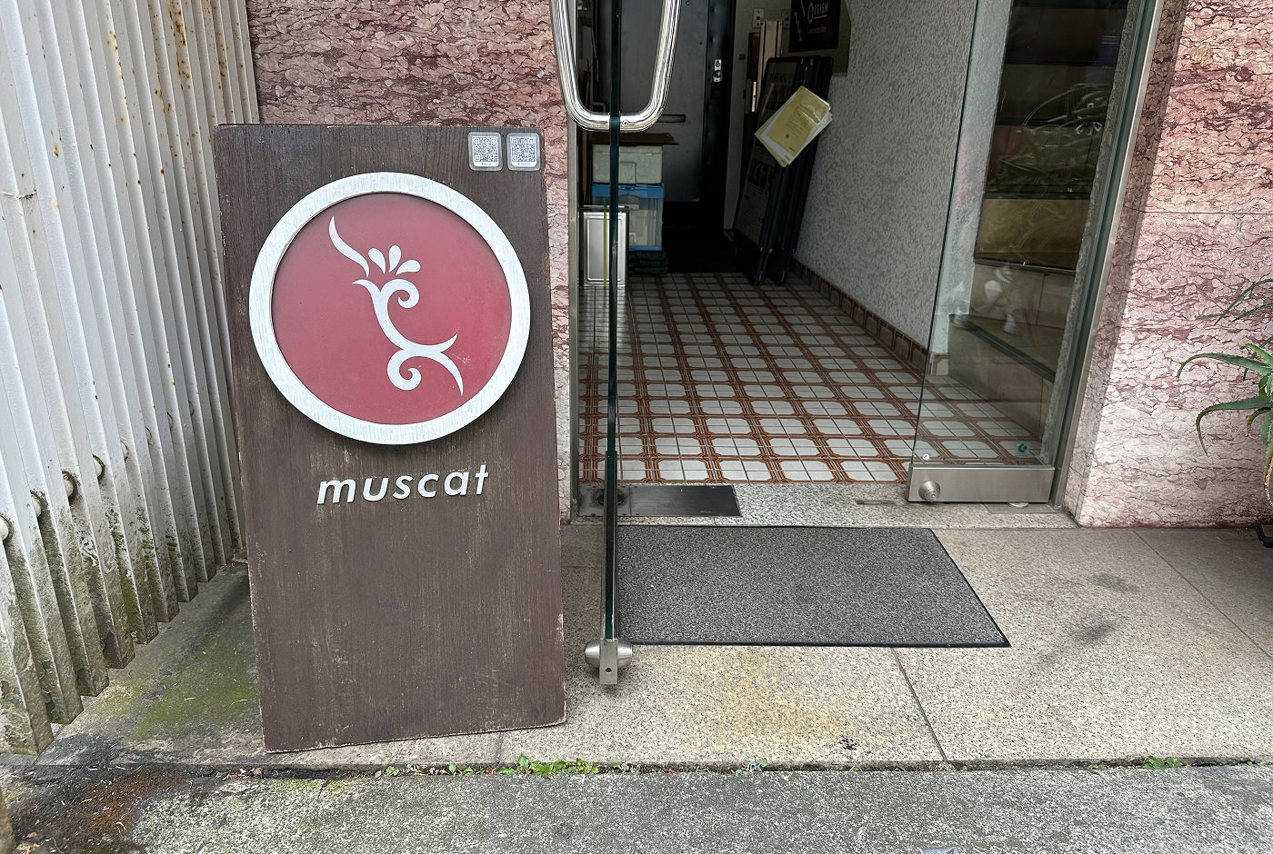 Ingång till ett hus med glasdörr. Till vänster står en brun träskylt med en logotyp i form av en röd cirkel med vit kant. Under lyder texten "Muscat".