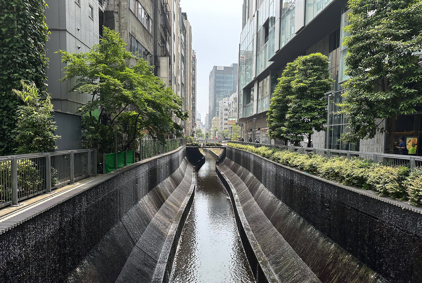 Smal kanal med vattenfall på båda sidorna ringlar sig genom en stad med höghus.