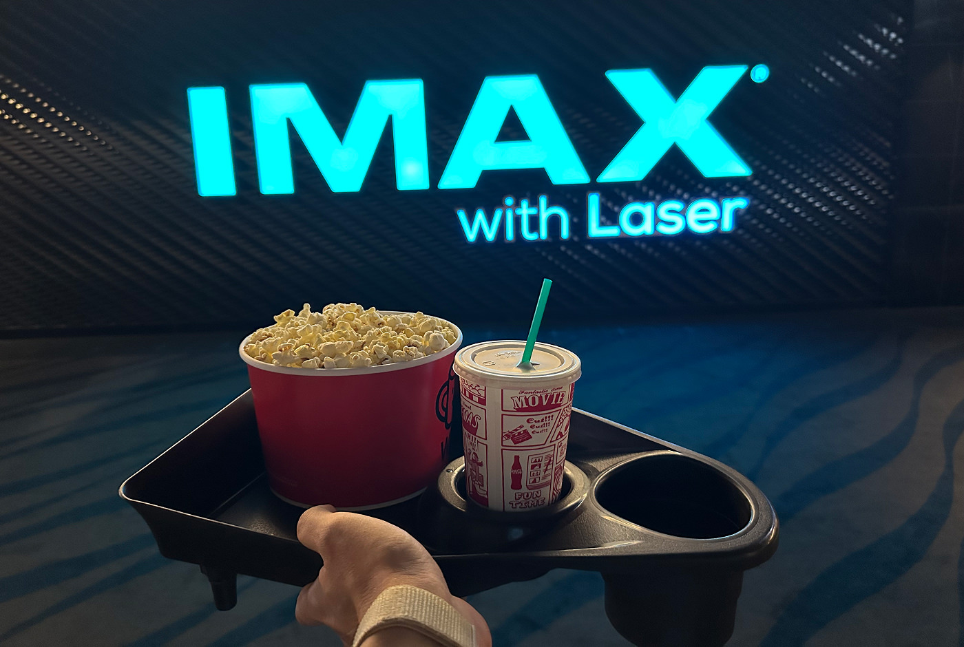 Hand håller i en svart bricka med en bytta popcorn och en pappersmugg med sugrör. På väggen i bakgrunden står det "IMAX with Laser".